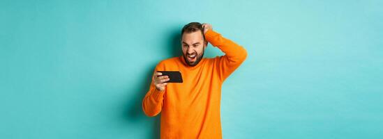 Image of man looking confused at mobile phone, watching something online, standing puzzled against light blue background photo
