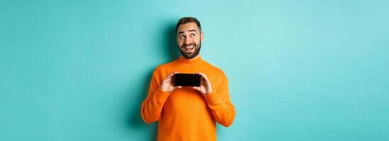 Online shopping. Young man thinking and showing smartphone screen, looking dreamy at upper left corner, standing over light blue background photo