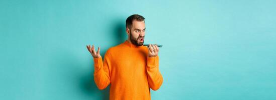 Angry man arguing on speakerphone, record voice message with mad face, standing over light blue background in orange sweater photo