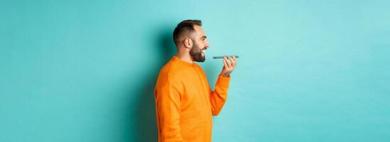 Profile of handsome bearded man recording voice message, holding mobile phone and talking on speakerphone, smiling happy, standing against turquoise background photo