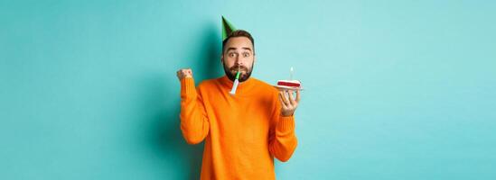Happy birthday guy celebrating, wearing party hat, blowing wistle and holding bday cake and doing fist pump excited, standing over turquoise background photo