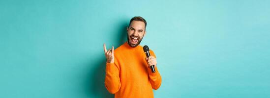 Joyful handsome man singing song in microphone, showing rock-n-roll gesture excited, standing over turquoise background photo