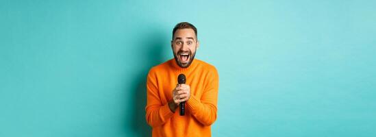 Handsome adult man perform song, singing into microphone, standing against turquoise background photo