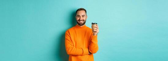 Handsome smiling man taking break, drinking coffee from takeaway, standing over blue background photo