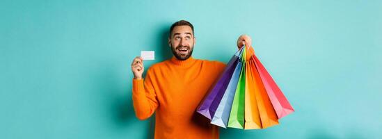 Happy aduly man showing credit card and shopping bags, standing against turquoise background photo