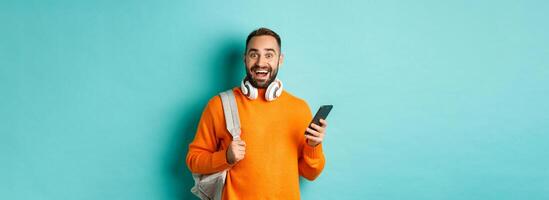Caucasian man with headphones and backpack staring at camera amazed after reading phone notification, standing over turquoise background photo