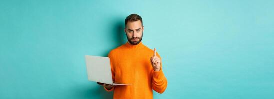 Image of adult man teaching online, showing finger while using laptop, explaining rules, standing over light blue background photo