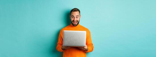 Excited man in orange sweater working on laptop, looking at computer screen amazed, standing over light blue background photo