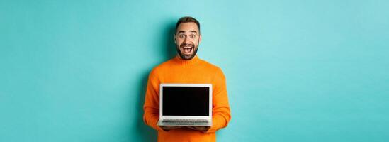 Handsome bearded man in orange sweater showing laptop screen, demonstrating promo, standing over light blue background photo