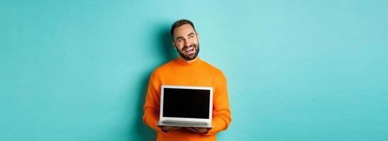 Happy caucasian man thinking and showing laptop screen, smiling pleased while pondering, standing in orange sweater against light blue background photo