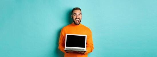 Amazed young man in orange sweater looking at upper left corner, showing laptop screen promo offer, standing over turquoise background photo