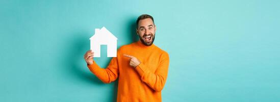 Real estate concept. Happy man pointing finger at paper home maket and smiling, standing over light blue background photo