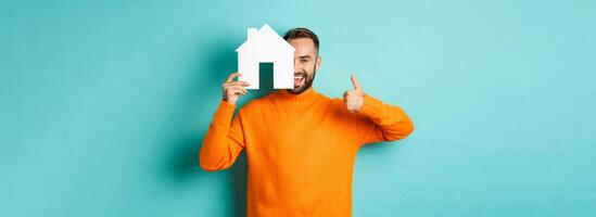 Real estate concept. Satisfied man recommending agency, showing thumb up and house maket, standing over blue background photo