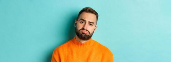 Close-up of upset young man sulking, looking disappointed and distressed, standing in orange sweater against turquoise background photo