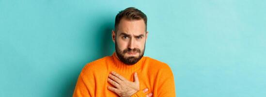 Close-up of shocked and startled man touching chesk, looking displeased and worried, standing against light blue background photo