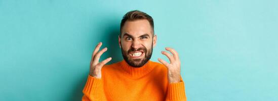Close-up of angry caucasian man looking mad, grimacing and shaking hands, having break down, standing over light blue background photo
