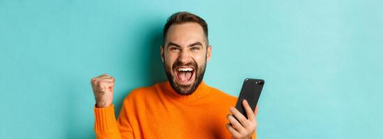 Image of man winning on mobile phone, making fist pump and rejoicing of victory, holding smartphone with satisfied face, turquoise background photo