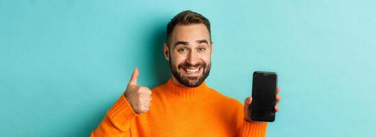 Close-up of attractive bearded man showing smartphone screen, thumbs up, recommending mobile app, standing satisfied over turquoise background photo