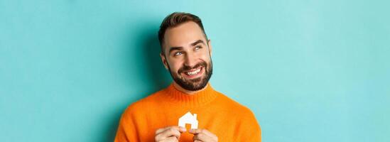 Real estate concept. Happy young man searching for home rent, holding house paper maket and smiling, standing over blue background photo
