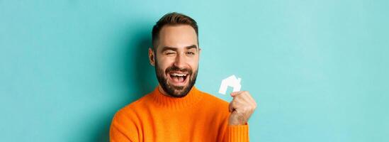 Real estate concept. Happy young man searching for home rent, holding house paper maket and smiling, standing over blue background photo