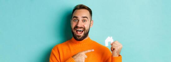 Real estate. Excited bearded man pointing at small paper house maket, standing amused over light blue background photo
