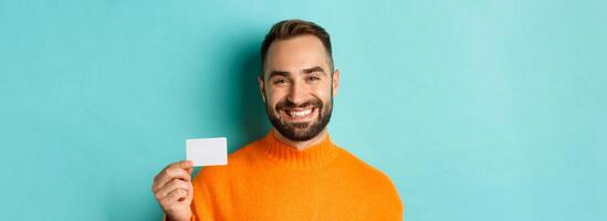 Close-up of handsome caucasian man going on shopping, showing credit card and smiling, standing over turquoise background photo