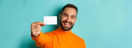 Close-up of handsome caucasian man going on shopping, showing credit card and smiling, standing over turquoise background photo