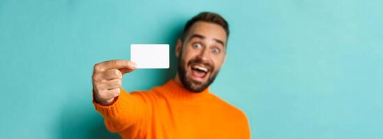 Close-up of handsome caucasian man going on shopping, showing credit card and smiling, standing over turquoise background photo