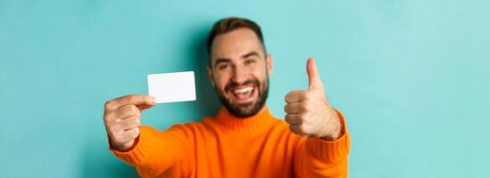Close-up of handsome caucasian man going on shopping, showing credit card and thumb up in approval, standing over turquoise background photo