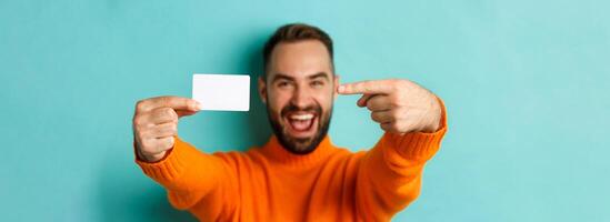 Close-up of handsome caucasian man going on shopping, showing credit card and smiling, standing over turquoise background photo