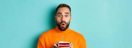 Close-up of happy adult man celebrating birthday, holding bday cake with candle and making wish, standing against turquoise background photo