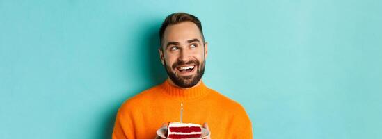 Close-up of happy adult man celebrating birthday, holding bday cake with candle and making wish, standing against turquoise background photo