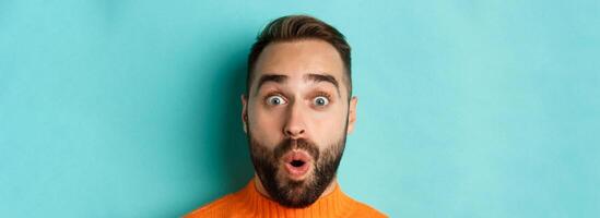 Headshot of handsome caucasian man with beard standing in orange sweater against turquoise background, saying wow and staring surprised at camera photo