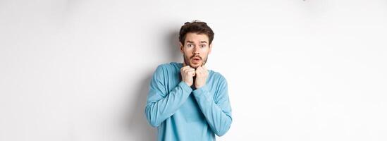 Image of scared young man trembling from fear, holding hands near face and staring at camera startled, standing over white background photo