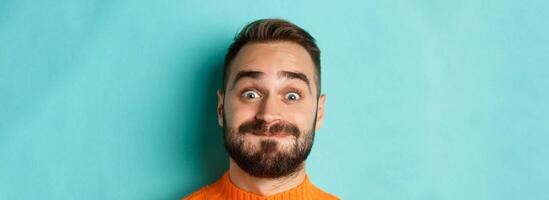 Headshot of funny bearded man showing faces and pouting, holding breath, standing in orange sweater against turquoise background photo