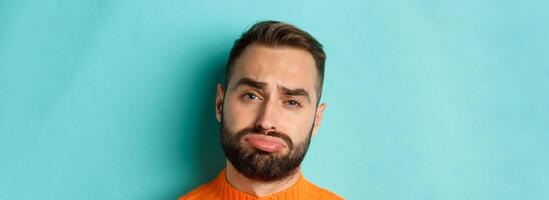 Headshot of sad and gloomy man complaining, pouting and frowning disappointed, standing against turquoise background photo