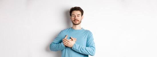 Portrait of young caucasian man with beard standing with hands on heart and closed eyes, dreaming about happy moments, standing over white background photo
