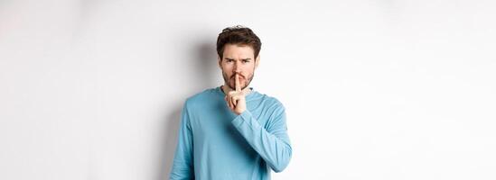 Angry man shushing at camera and telling to be quiet, frowning upset, standing over white background with finger on lips, taboo gesture photo