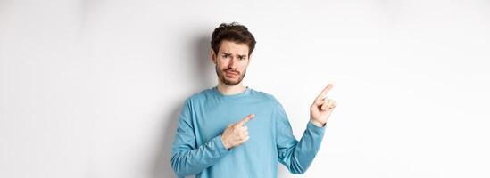 Skeptical young man with beard grimacing unsatisfied, frowning and pointing at upper left corner logo doubtful, standing upset over white background photo