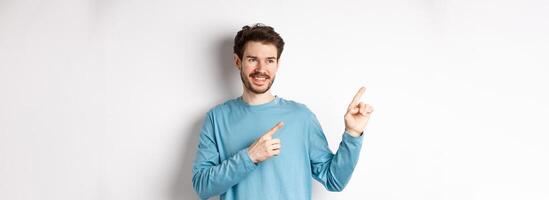 Portrait of young modern man in blue sweatshirt pointing fingers at upper left corner and looking away, smiling pleased as showing advertisement on white background photo