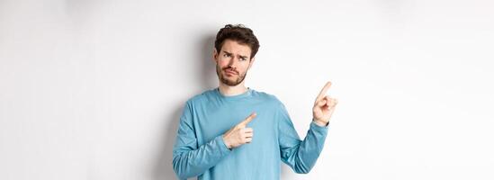 Skeptical and unimpressed young man grimacing, pointing fingers and looking at bad promotion, standing dissatisfied over white background photo