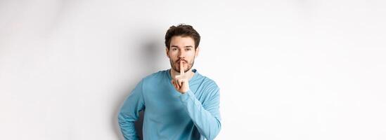 Attractive bearded man asking to keep quiet, showing taboo hush gesture and looking at camera calm, standing over white background photo