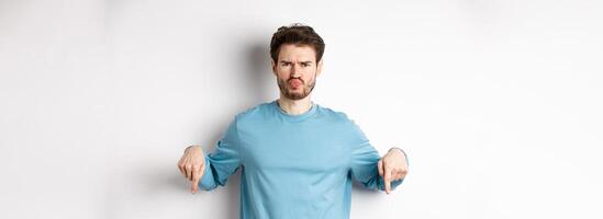 Sulking man pointing fingers down at something bad, frowning and complaining, standing over white background photo