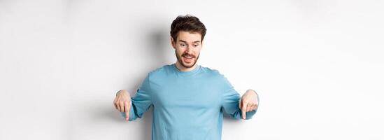 Excited and happy young man looking and pointing down at awesome deal, checking out promotion offer, standing on white background photo
