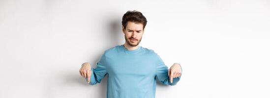Skeptical boy pointing fingers down and frowning disappointed, looking at bad promo offer, standing on white background photo