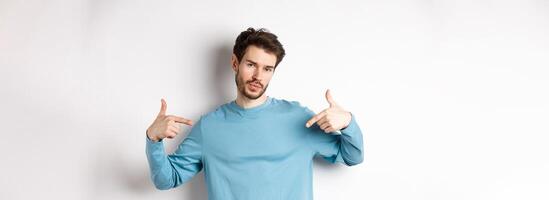 Young man with beard feeling confident, pointing at himself and making smug face, standing on white background cool and sassy photo