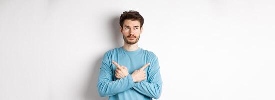 Indecisive man making choice, pointing fingers sideways at two variants and deciding, looking pensive at left side, standing on white background photo