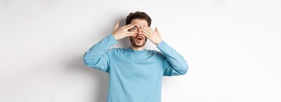 Amazed smiling man covering eyes with hands but peeking through fingers at something awesome, standing over white background photo