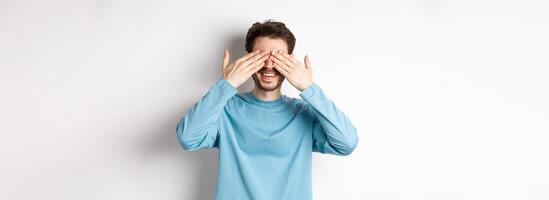 Smiling caucasian man waiting for surprise with closed eyes, expect gift, standing over white background. Celebration and holidays concept photo