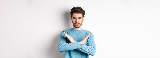 Young man with beard looking serious, making cross gesture to stop something, prohibit action, standing against white background photo